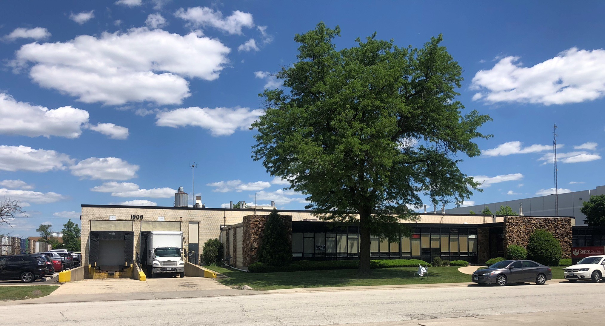 Street view of tan Industrial warehouse with two loading docks and a large oak tree, Cawley CRE