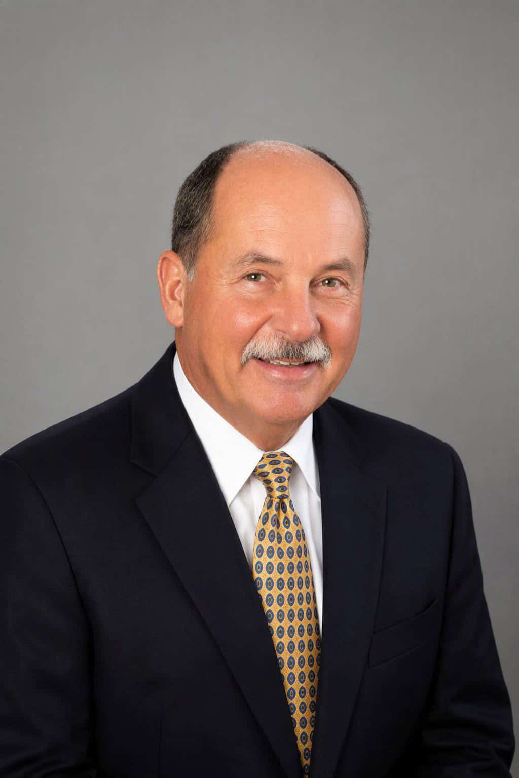 Professional portrait of Duke Botthof in a suit and tie against a gray background.
