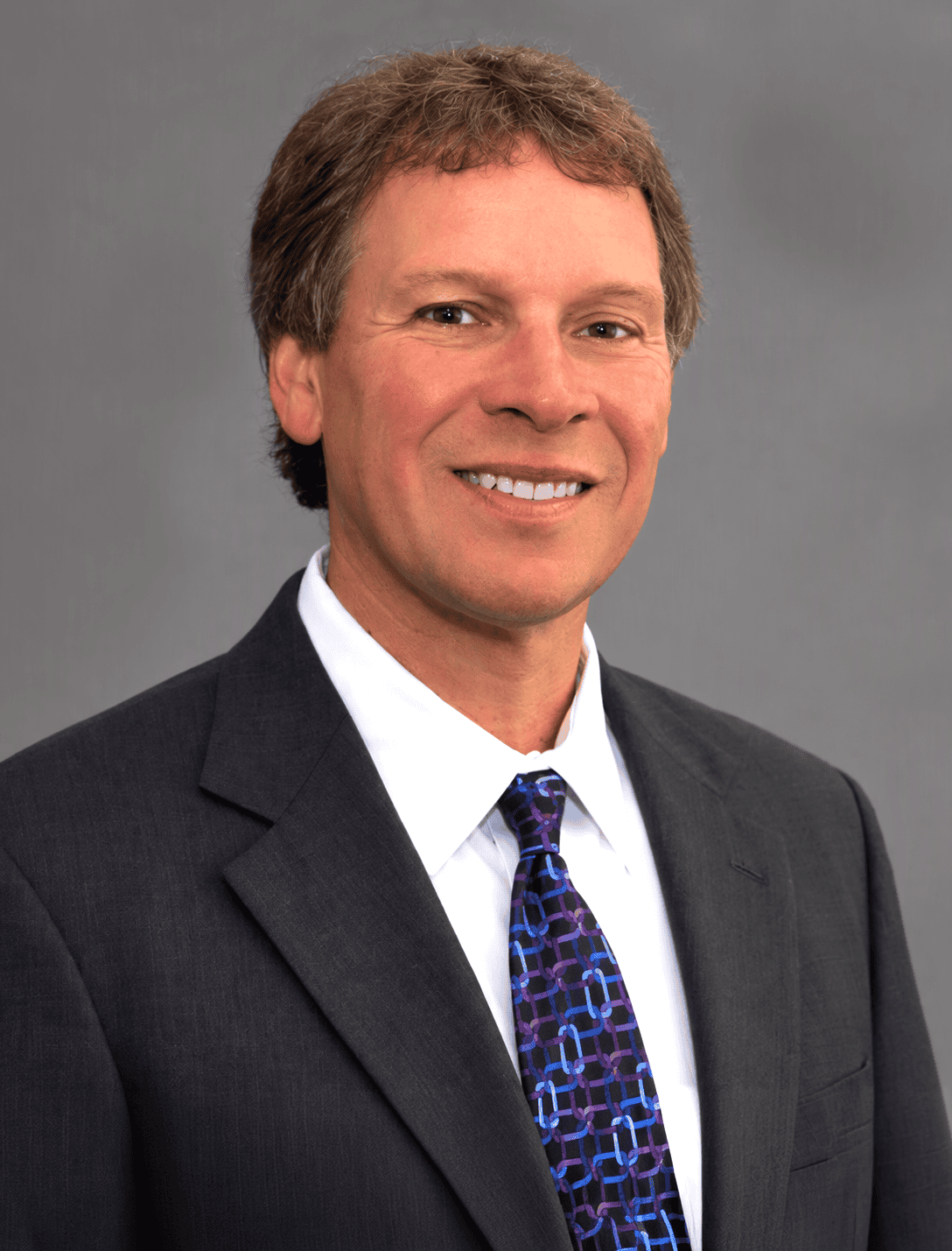 Professional portrait of a man in a suit and tie, smiling, against a gray background.
